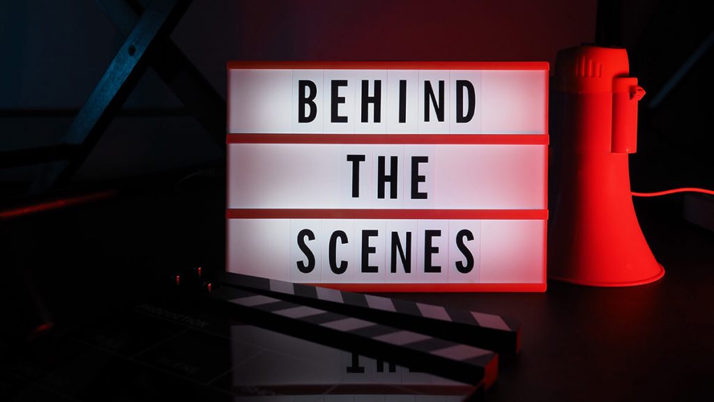 Tafel mit schwarzen Buchstaben auf weißem Hintergrund mit "Behind the scenes" und rotem Megafon rechts