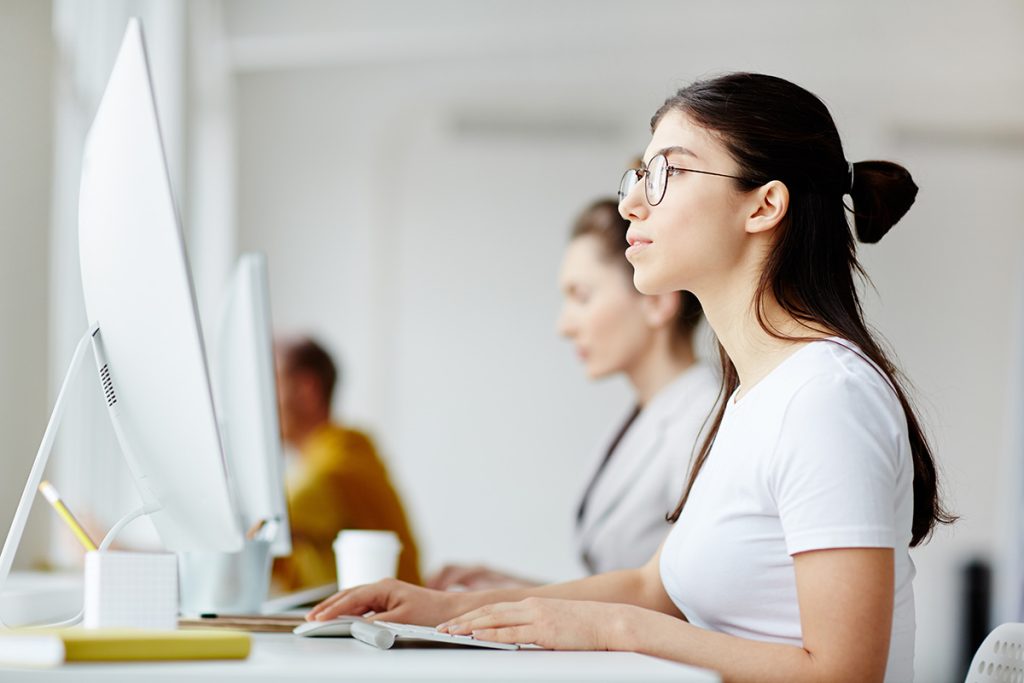 Junge Frau mit Brille in weißem Shirt sitzt vor Computer mit weißem Bildschirm